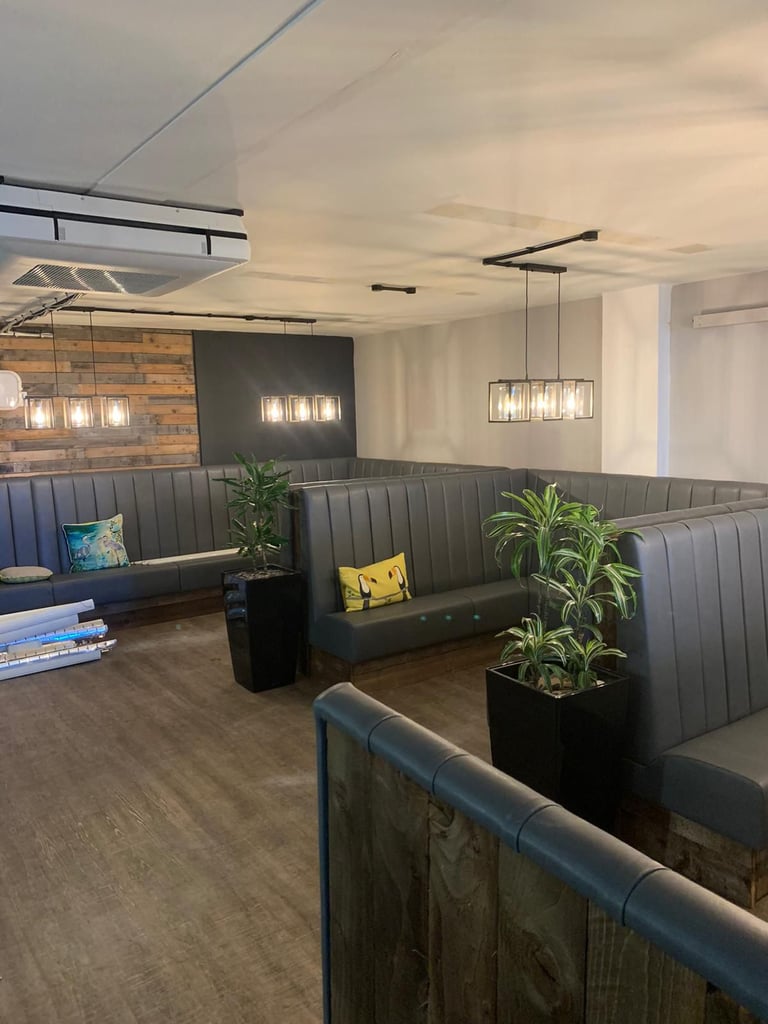 Modern restaurant interior with gray banquette seating, exposed brick wall, pendant lighting, potted plants, and wood flooring